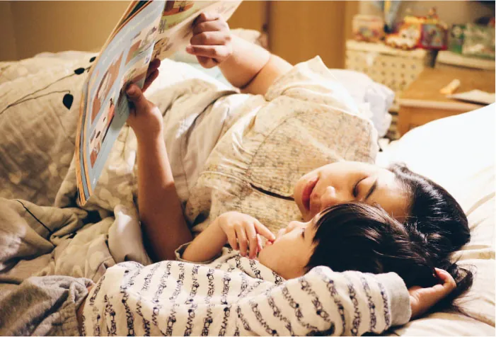 Photo of adult and child laying on a bed reading a book together.