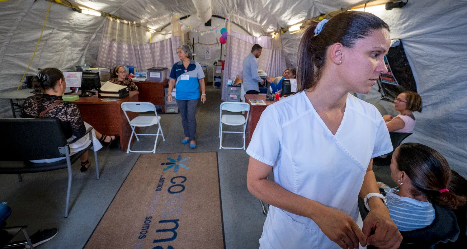 In a large tent with desks and chairs, health care professionals walk and sit with clients.