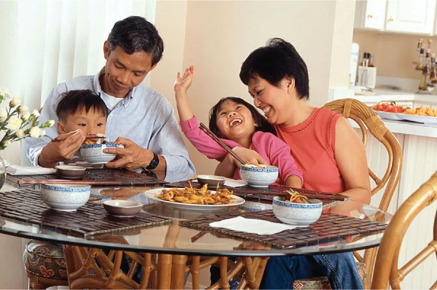 Image of a family seated around a table eating a meal with chopsticks. An adult male is feeding a boy on his lap. An adult female has a girl on her lap.