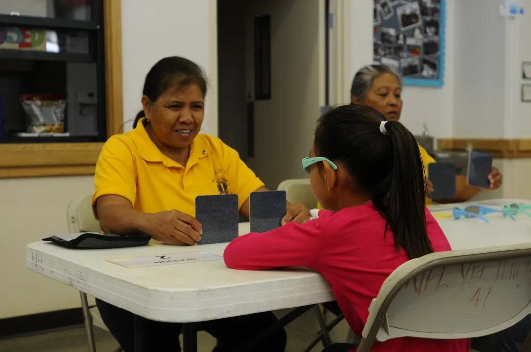 A school nurse sits across a table facing a young student. The nurse holds up two black squares on the table.