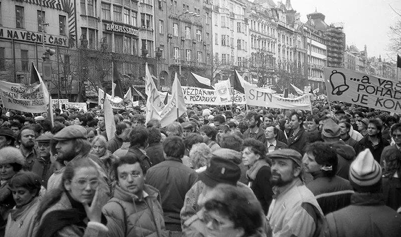 Protesters crowd a street lined with old European buildings. Several protesters hold up banners.