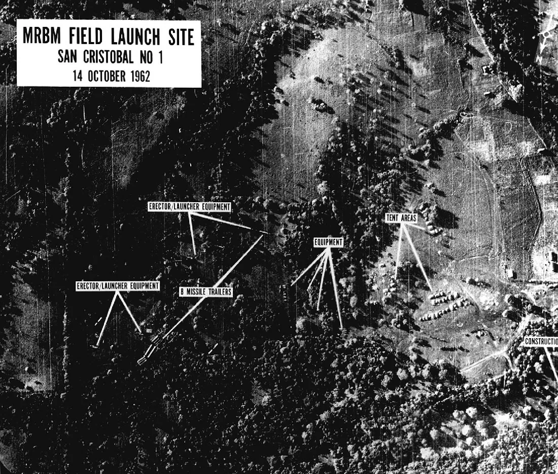 An aerial terrain photograph is shown. Most of the left side of the picture shows trees from above, with areas within the trees labeled “Erector/Launcher Equipment” and “B Missile Trailers.” In the middle of the photograph there are four small shapes labeled “Equipment.” In the middle right of the photo there are two areas with small bundles shown in rows that are labeled “Tent Areas.” The bottom right of the photograph has a label with “Construction.” A label in the top right indicates: MRBM Field Launch Site, San Cristobal NO 1, October 14, 1962.