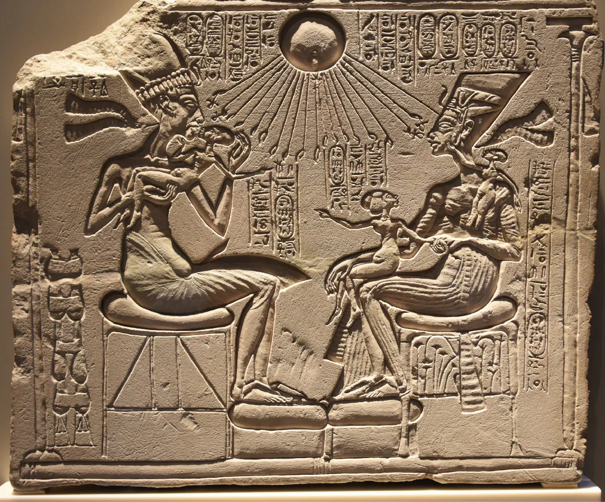 A picture of a brown stone carving is shown with a broken corner at the top left. The carving shows an orb in the middle at the top with rays coming out and symbols at the end of the rays. Hieroglyphics are carved across the top of the stone and below the rays from the orb as well as along the right side of the carving. On the left side of the carving a man sits on a padded stool with his bare feet on a padded footrest. He wears a tall detailed headdress and has a cloth around his waist. He holds a small person in his arms and is kissing them. In the right half of the carving a woman is seated on a padded stool with her bare feet on a smaller padded footrest. She wears a decorated headdress and long cloths on her body. She holds a small person on her lap and another is on her shoulder. Obscure designs run along the bottom left of the stone and the edges of the carving have decorative lines all around.