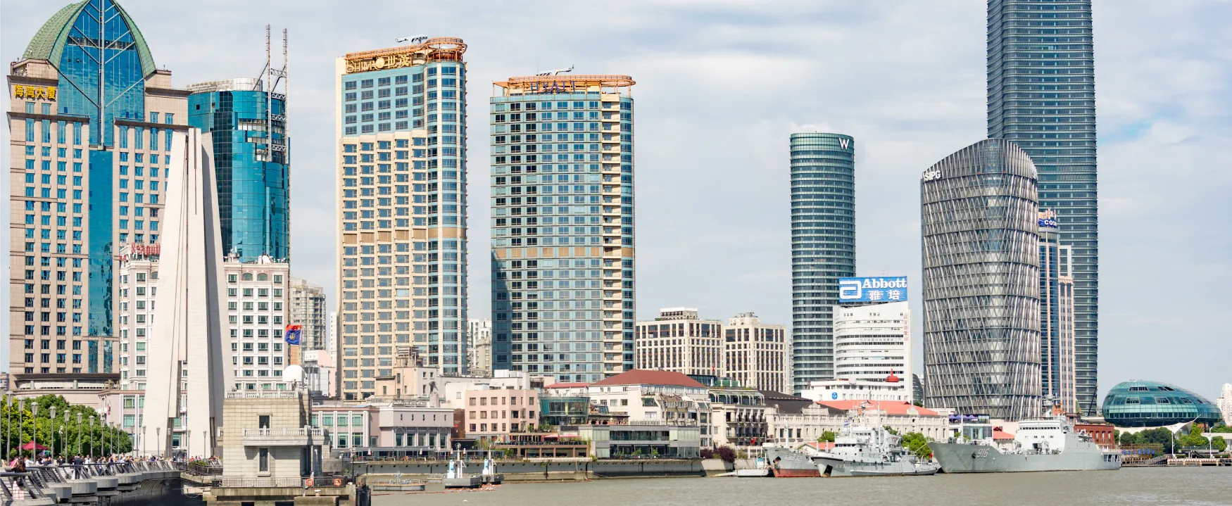 A dynamic urban panorama showcasing modern skyscrapers towering over a bustling waterfront. Ships are docked along the river, while people stroll on a promenade against a partly cloudy sky, reflecting a vibrant city scene.