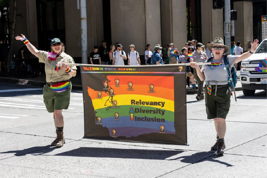 Two people walk down a street carrying a parade banner between them. The banner has the shape of Washington state on it, presented in rainbow colors. Inside the shape are the words “Relevancy, diversity, inclusion”.