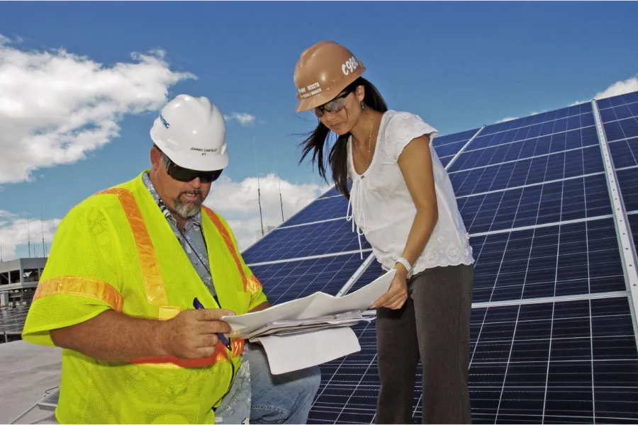 Photo of two individuals wearing hard hats, reviewing paperwork, with solar panels in the background.