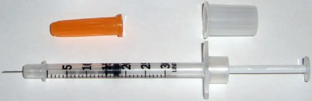 An open insulin syringe with a needle lays in front of two vials of insulin. An orange cap is between the syringe and the vials.