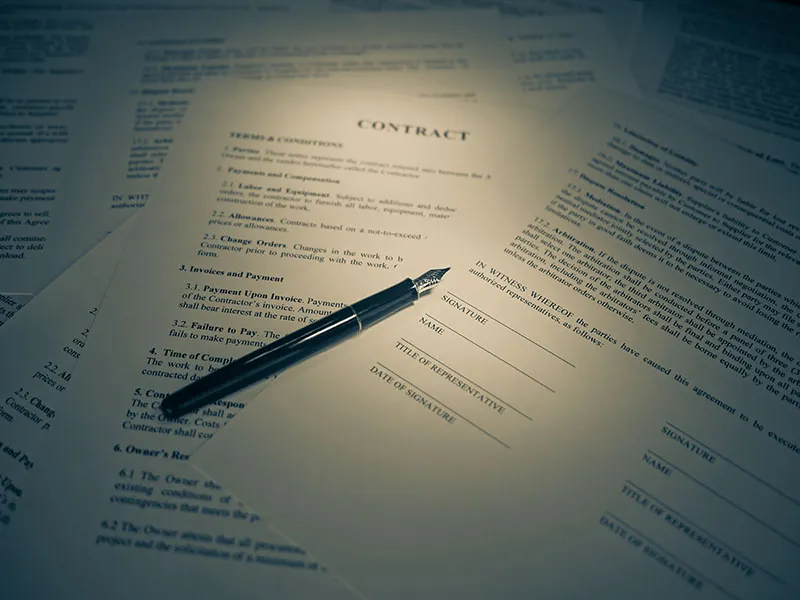 A stack of papers is loosely spread out on a table. The word contract appears in the top center of one page, and a pen rests on top of the page.