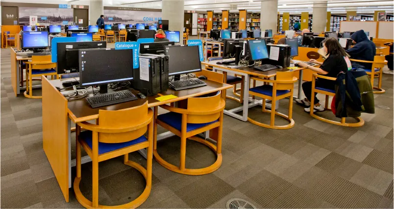 A library is shown with several tables and chairs with desktop computers on the tables. People are seen working on the computers.