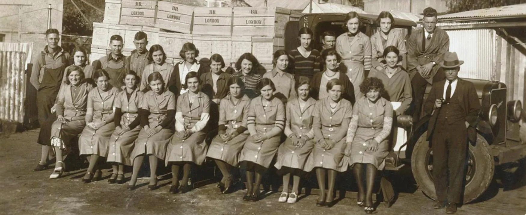 A large group of factory or distribution workers, mostly young women, pose for a vintage photo outside with a truck and crates marked 'BR LONDON' in the background.