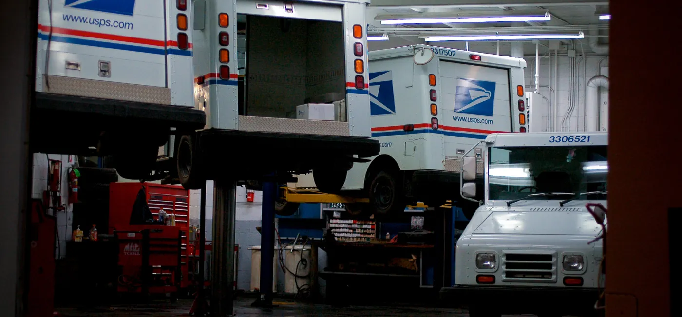 This is a photo of a car mechanic’s shop. There are three United States Postal Services trucks being serviced, and one not being serviced.