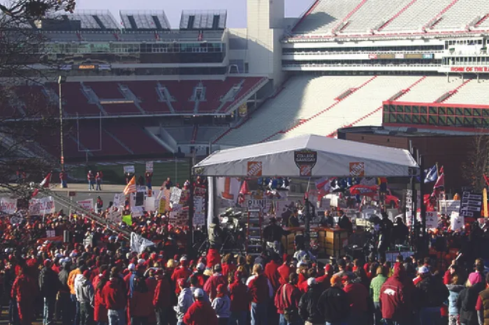 A photograph shows a crowd of people standing near a stadium at a tent.