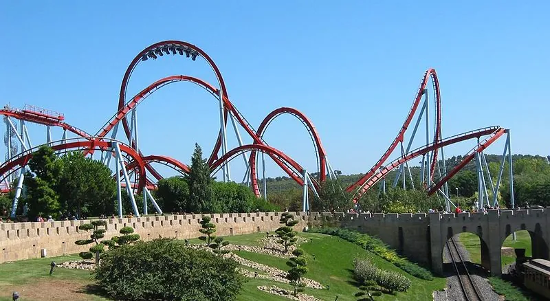 In this figure the Dragon Khan rollercoaster in Spain’s Universal Port Aventura Amusement Park is shown. There are mostly curved paths in the rollercoaster. Near to the rollercoaster there is the track of rollercoaster cart under a bridge. There are some trees near the track.