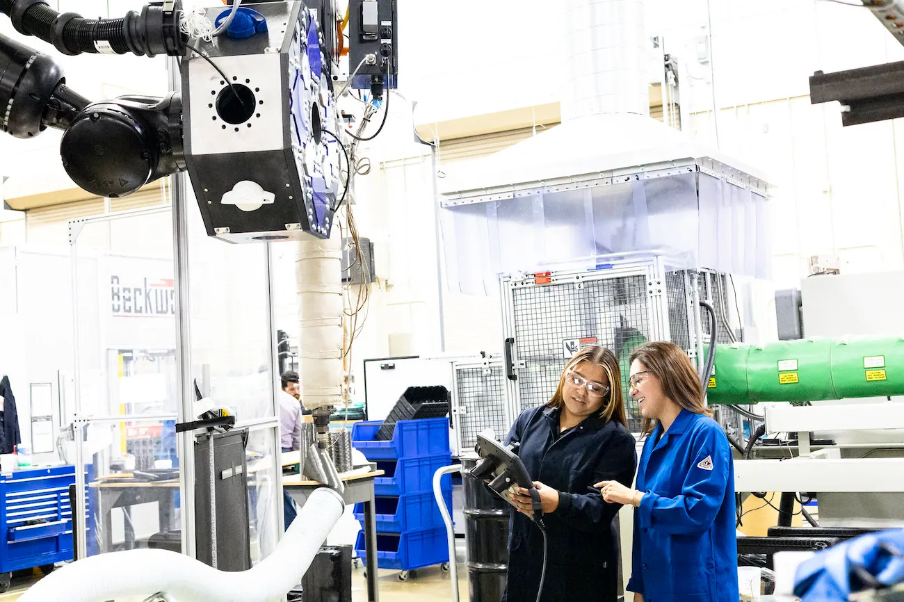 Two people inside a manufacturing facility look at a tablet or similar instrument. They are surrounded by various types of equipment, materials, and storage.