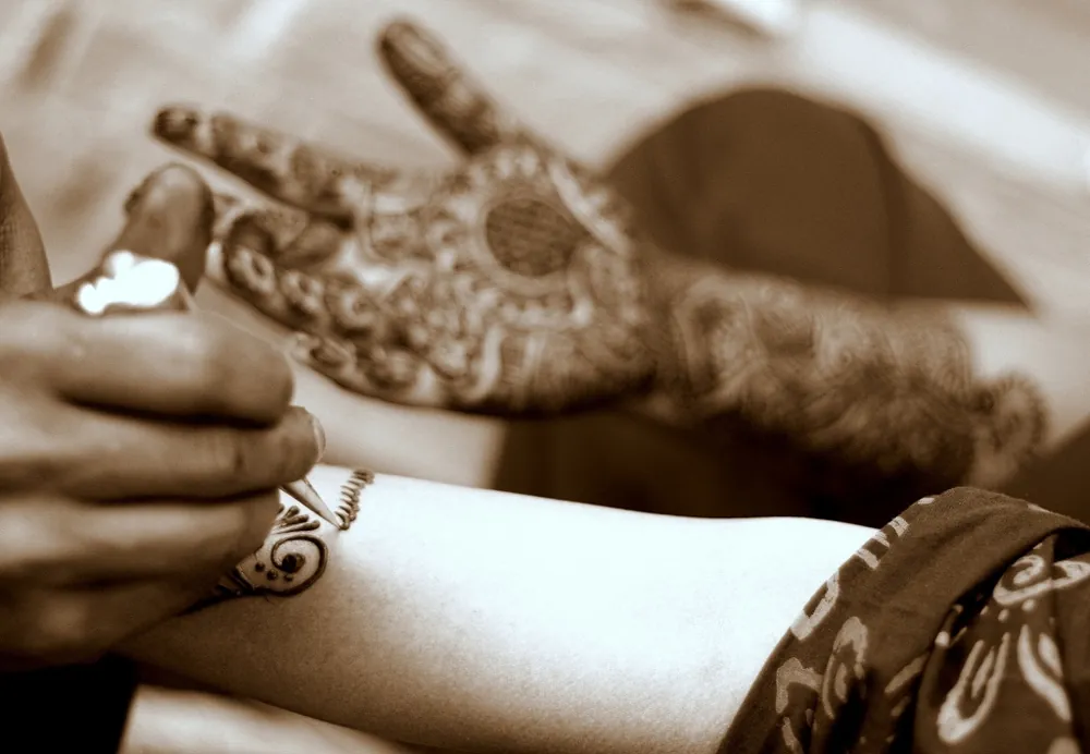 The photo shows a woman’s hands being covered in intricate henna designs.