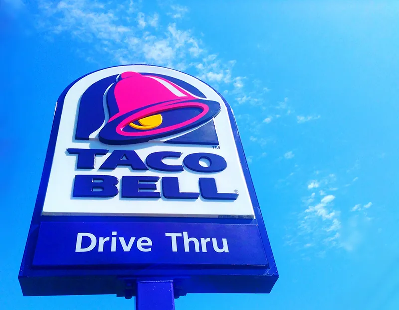 A large taco bell drive through sign is seen with blue sky and clouds behind it. At the top of the sign is a bell, with the words Taco Bell Drive Thru underneath.
