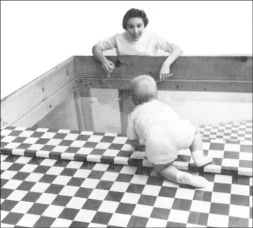 Photo of infant on checkered table, stopping before glass portion, while mother encourages child to come to her.