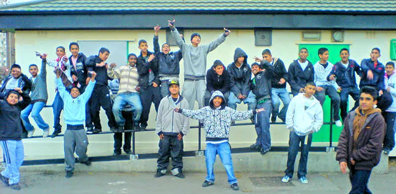 A group of about 25 young adults pose, some sitting or standing on a railing and some in front of it, on a sidewalk near a paved road.