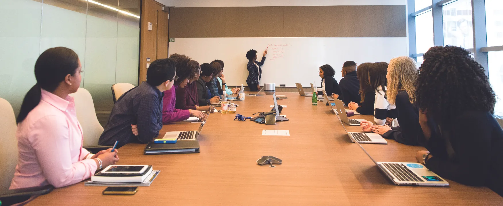 A photograph shows a group of people with laptops and notebooks sitting around a large conference table and looking up at someone writing on a board.