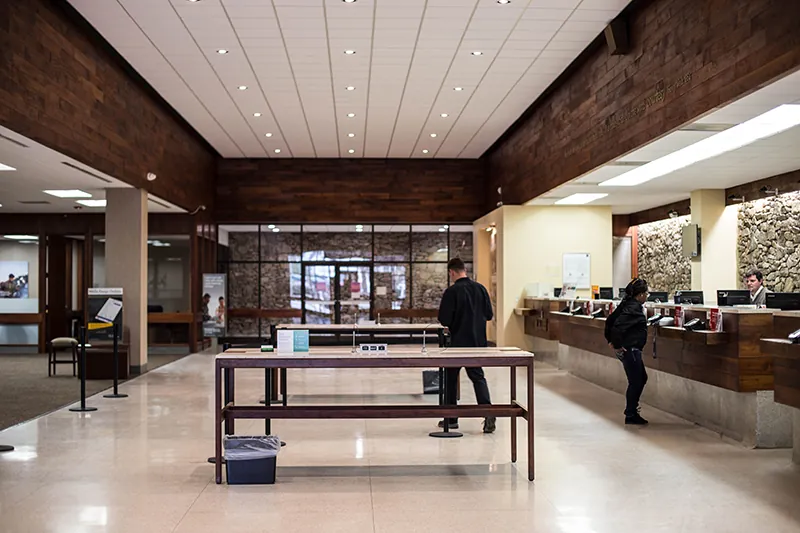 An photo shows the inside view of an office reception area. A few people can be seen standing in front of a counter with multiple computers and telephone systems place on it. 2 large tables are also occupying the area in front of the reception counter.