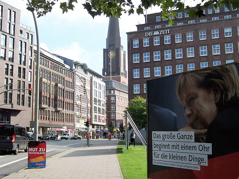 A large political poster for German Chancellor Angela Merkel stands alongside a wide sidewalk on a city street in Germany.