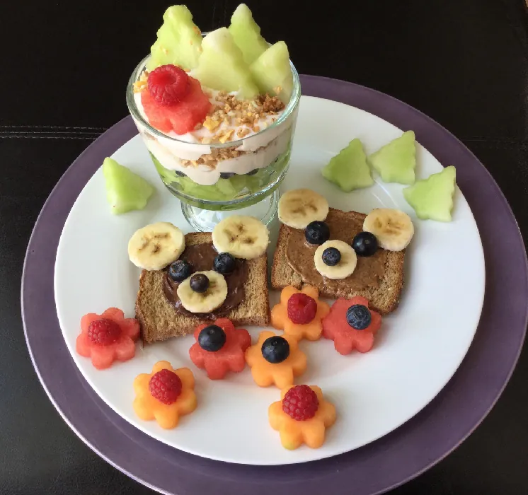 A dessert cup filled with yogurt, granola, and honeydew, with the melon cut into the shape of evergreen trees. A plate contains flower-shaped pieces of cantaloupe and watermelon that have a single raspberry or blueberry as the flower’s center. Two slices of whole grain toast have a circle of chocolate spread to make a teddy bear face, using banana slices for ears and a nose and blueberries for the eyes.
