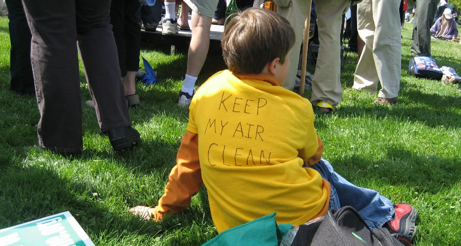 A child sits on the grass at an environmental rally wearing a shirt that says Keep My Air Clean.
