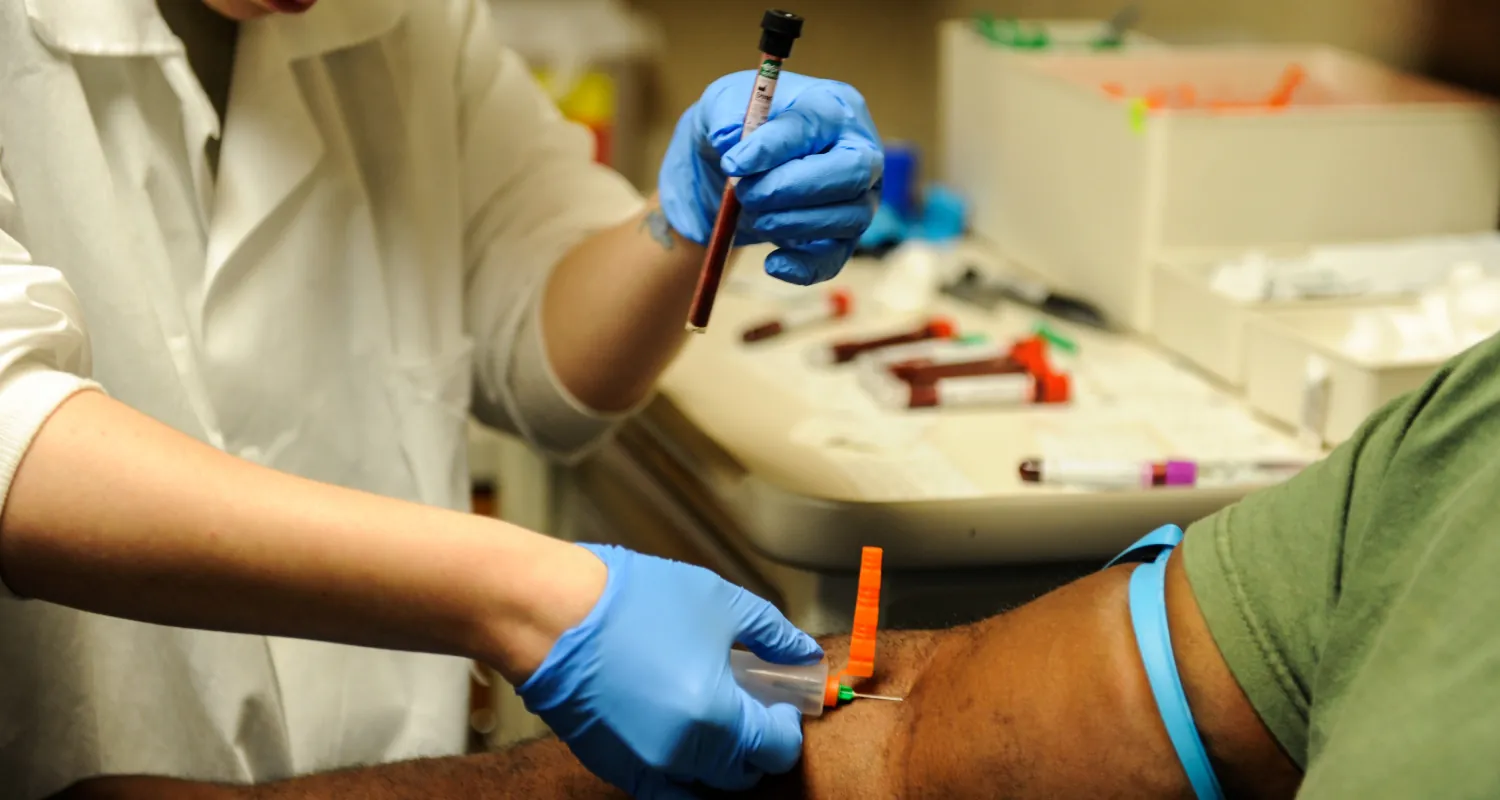 A healthcare worker, wearing blue gloves, extracts a blood sample from a person's arm using a syringe and test tube in a medical setting. 