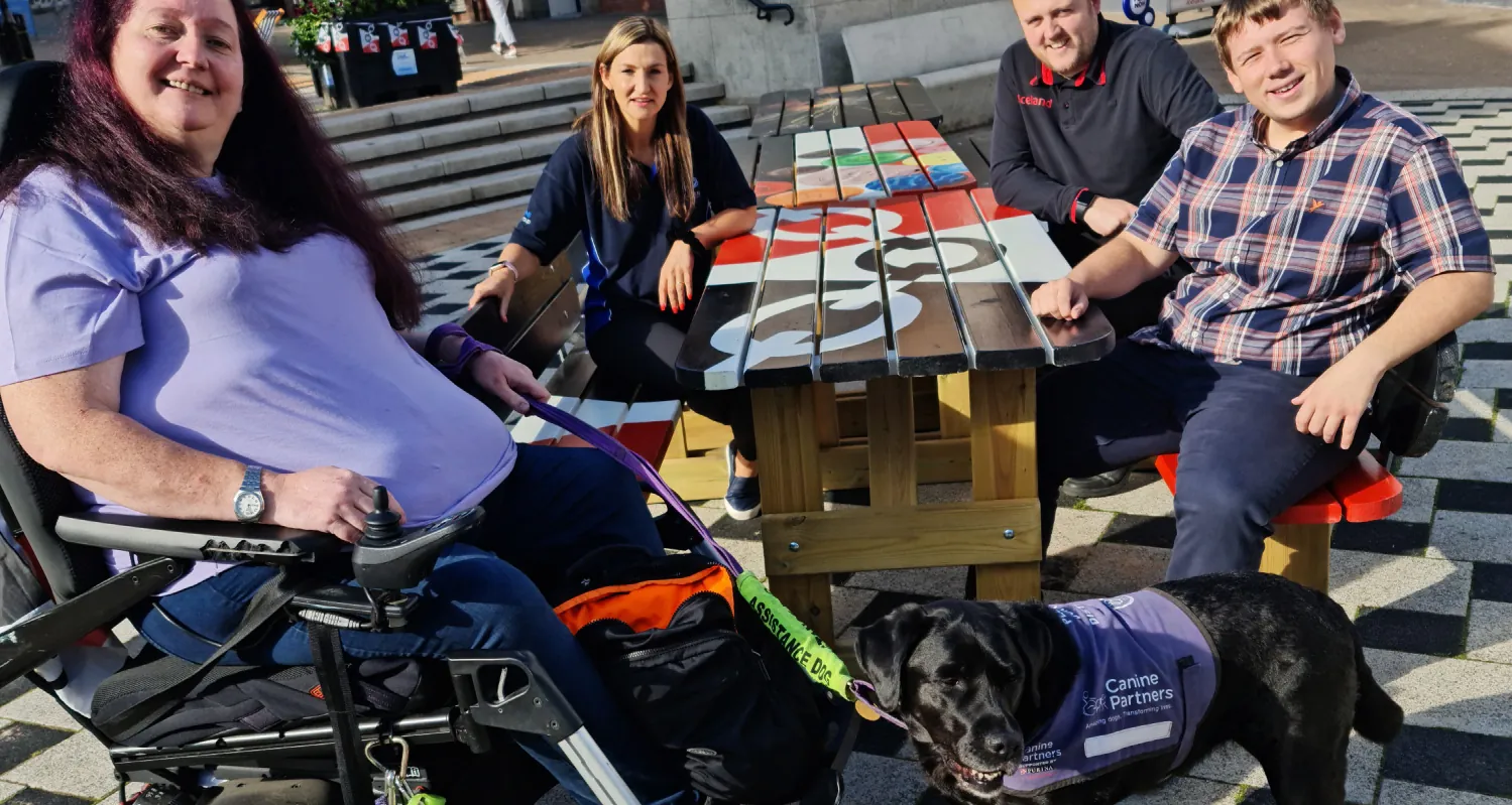 A photograph of an elderly woman sitting in a wheelchair and being assisted by a service dog. Three other individuals are in the picture sitting at a painted picnic table.