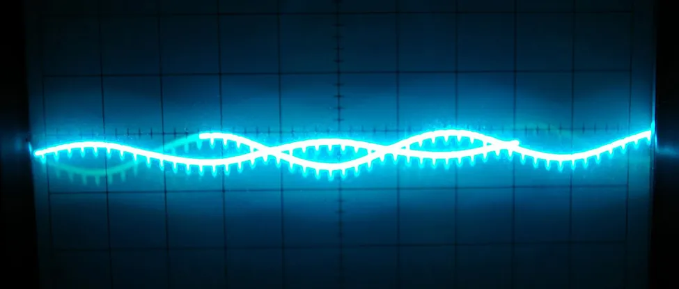 A close-up shot of an oscilloscope screen displaying a vibrant neon blue waveform. The waveform is complex, featuring a main undulating curve with smaller, uniform ripples or serrations along its entire length. There appears to be a second, fainter waveform of a similar pattern slightly offset and overlapping, creating an intricate, almost braided visual effect. The background is a dark blue grid, typical of an oscilloscope display, with lighter grid lines visible through the glowing waveform, indicating precise measurements.