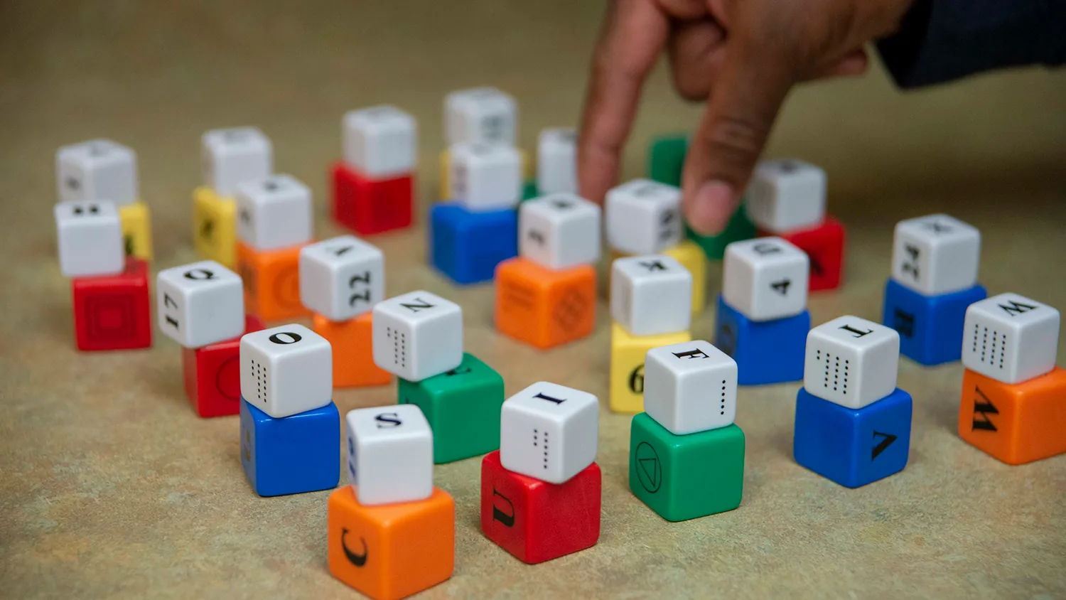 A collection of colored blocks with various symbols, letters, and numbers, with some blocks on top of others and a hand moving a pair of blocks. 