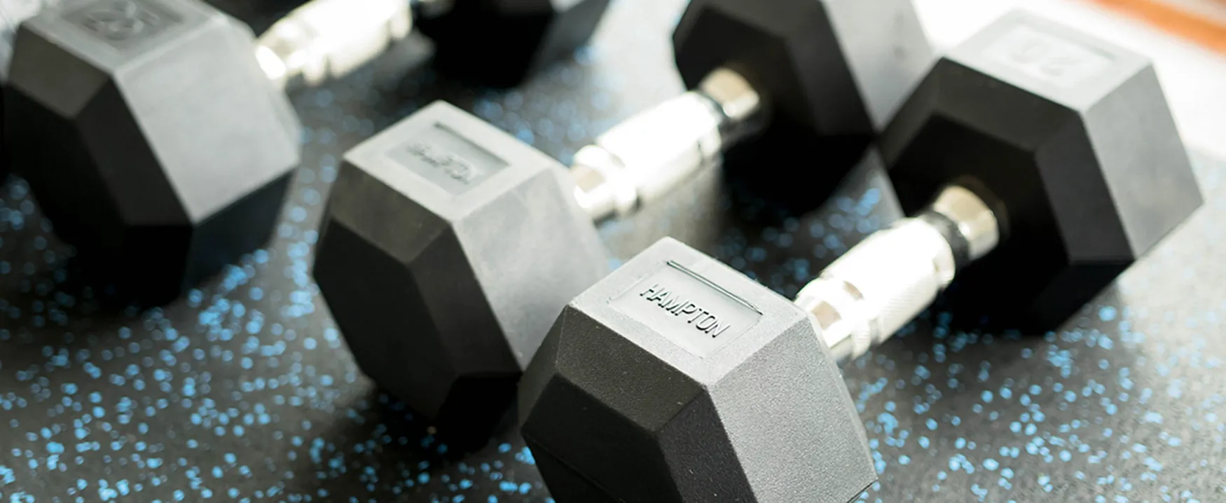 Close-up of several black hexagonal dumbbells with chrome handles, some showing 'HAMPTON' and '20,' resting on a black and blue speckled gym floor, ready for a workout session.