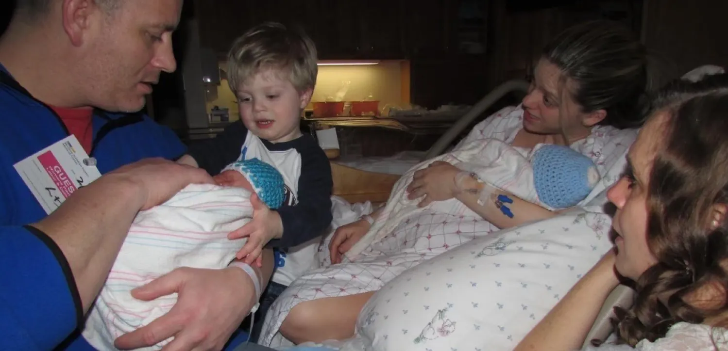 Parents introduce their young son to his newborn twin brothers in a postpartum hospital room. A support person looks on.