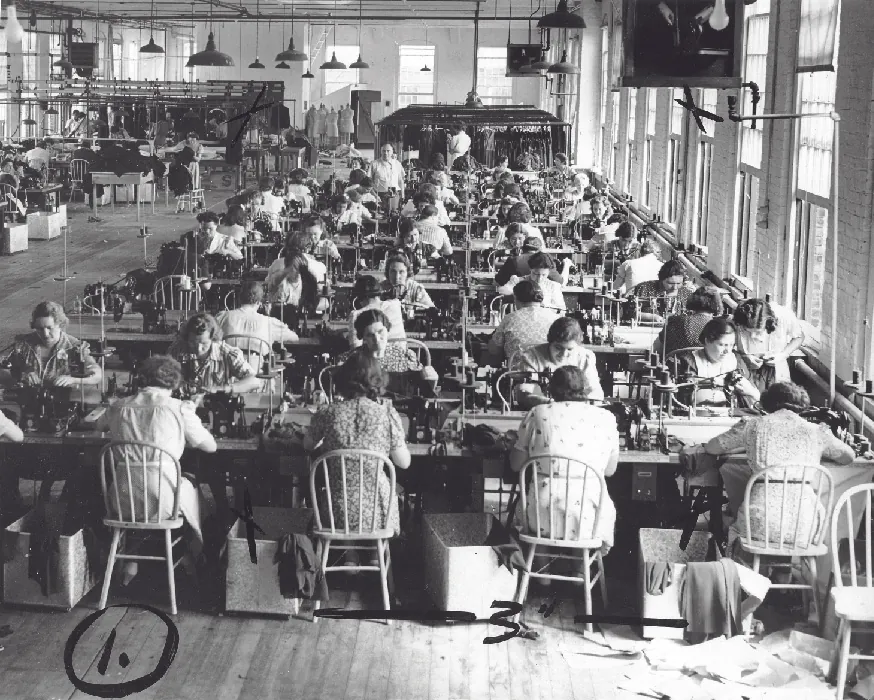 Workers sit sewing at both sides of long tables in a large room. Tall, open windows line the brick walls of the building.
