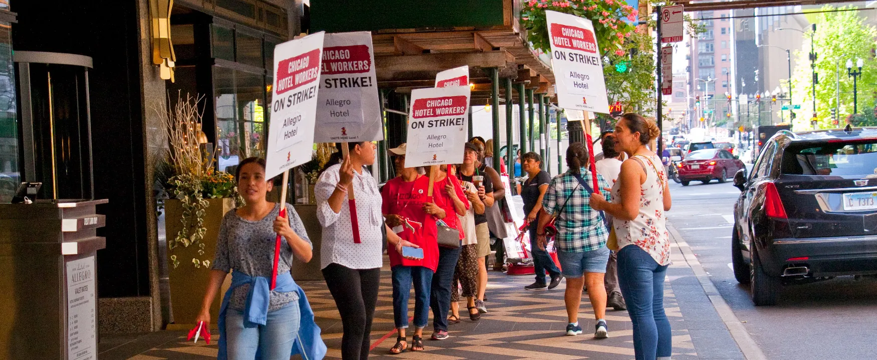 Chicago hotel workers picket outside the Allegro Hotel, holding signs that read 'CHICAGO HOTEL WORKERS ON STRIKE!' as traffic passes by on the busy city street.