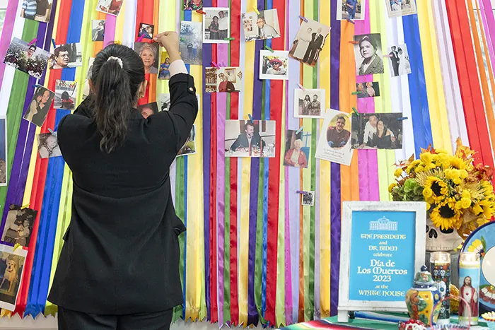 A person with long hair tied back in a ponytail, dressed in a black outfit, is pinning a photo to a colorful backdrop made of vertical ribbons in red, orange, yellow, green, blue, and purple. The backdrop is adorned with various photographs. In the foreground, there is a table with a sign that reads 'Día de los Muertos 2023 White House,' surrounded by sunflowers, candles, and other decorative items.