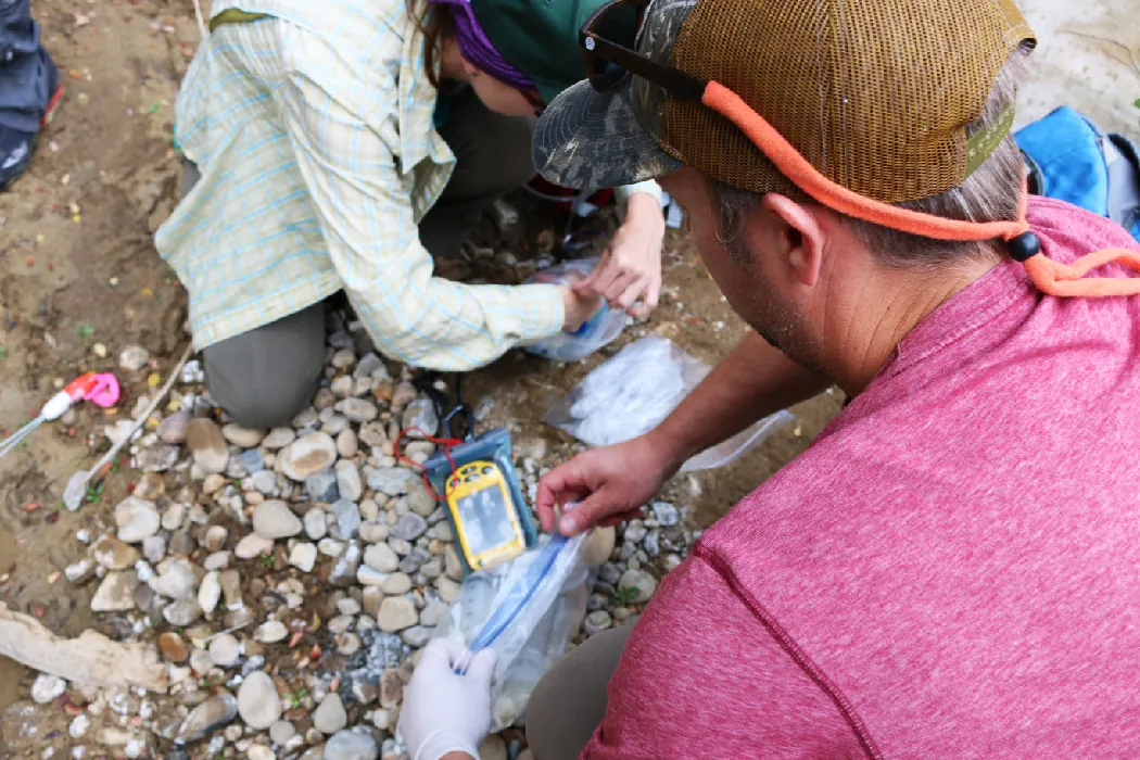 Two researchers kneel in a rocky shallow stream and collect water in zip lock bags.