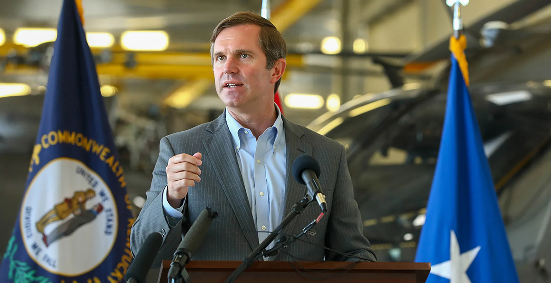 Governor Andy Beshear stands at a podium with various flags surrounding him.
