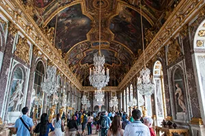 The Hall of Mirrors at Versailles features an al fresco and gilded domed ceiling; many large, hanging crystal chandeliers; exquisite painted panels on the walls; and mirrors—lots and lots of mirrors—to give an illusion of space. Tourists are pictured roaming the Hall.