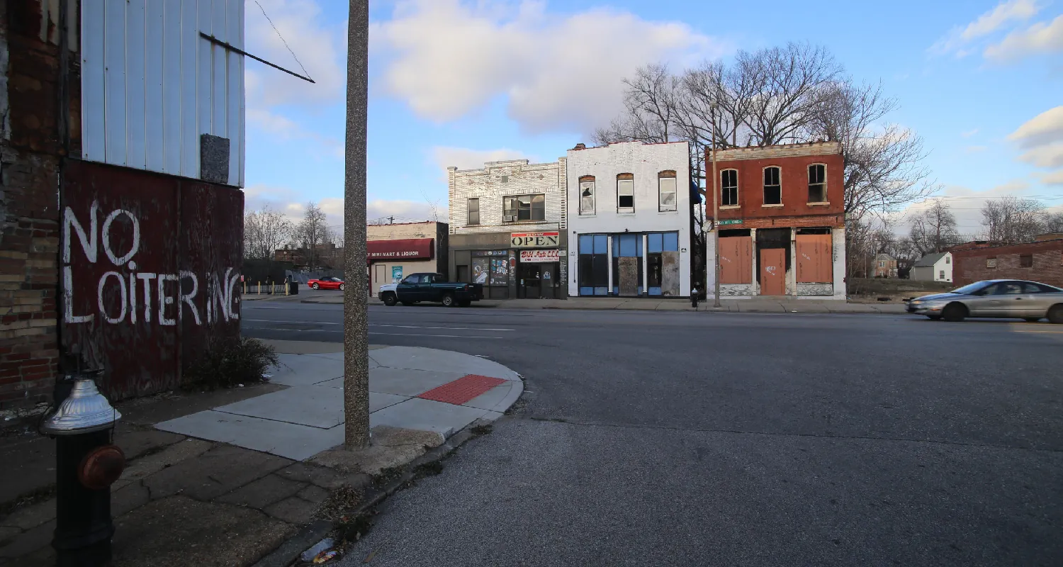 A building on a street corner has “no loitering” spray painted on the side. Across the street are four storefronts, two of which have boarded up windows.