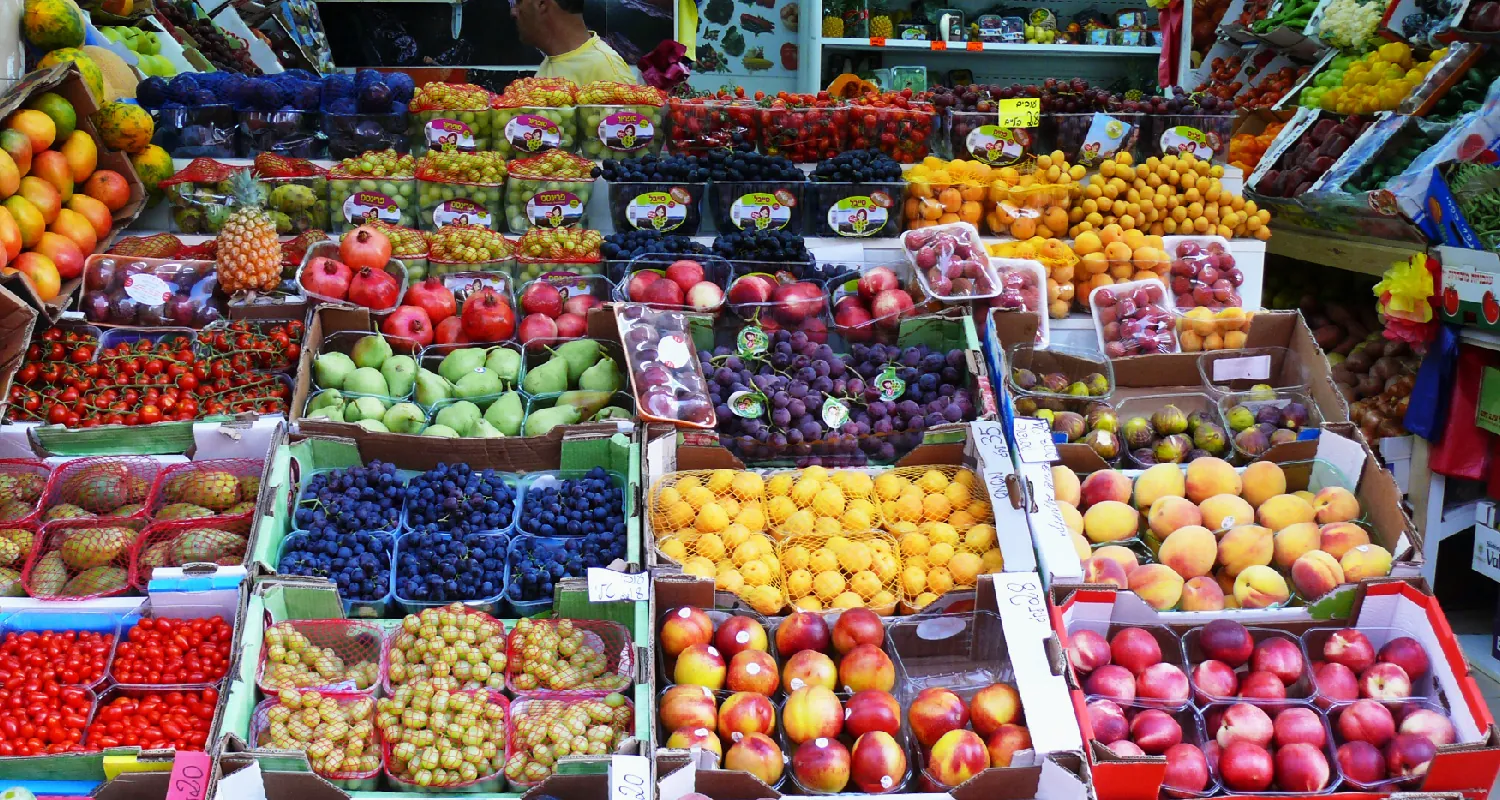 A market has colorful fruits and vegetables in boxes on a table.  The fruits and vegetables include plums, peaches, Brussel sprouts, tomatoes, blueberries, lemons, pears, pineapples, and mangos.