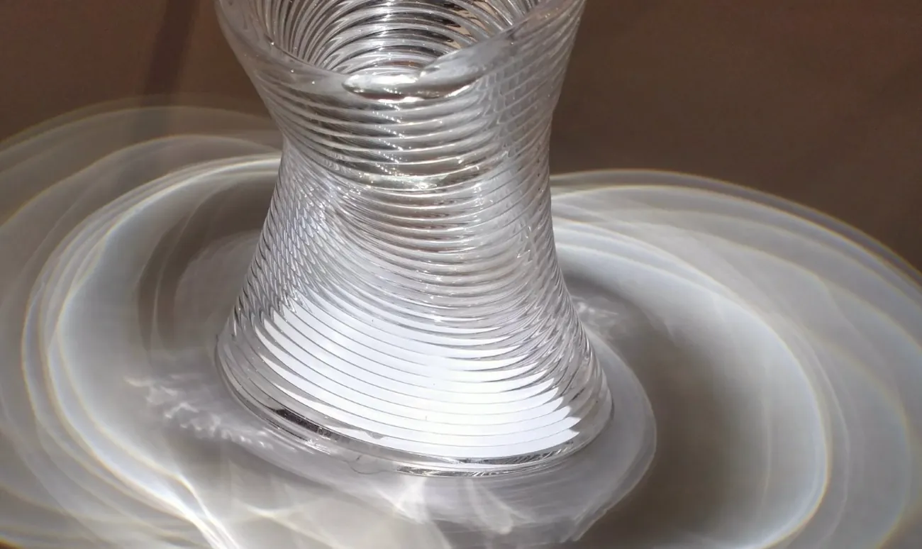 An artistic piece of glass in an irregular, roughly cylindrical shape. It is wider at the base, narrows somewhat toward the middle, and widens again at the top. Bands circle it. It is lit in such a way to produce intersecting circles of light on the display surface.