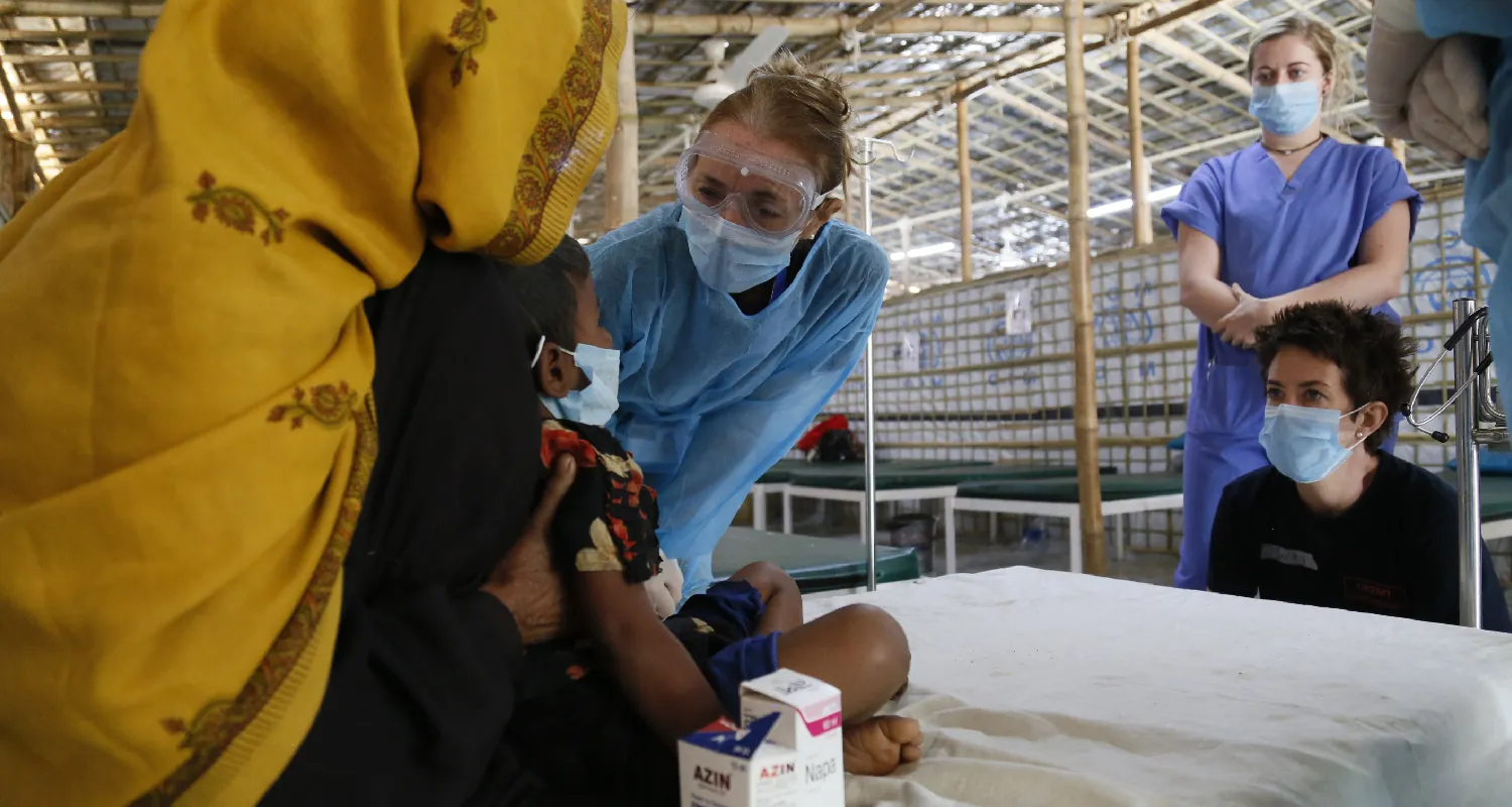 An adult draped in a headscarf sits with a child on a hospital bed in a temporary clinic. Four medical personal wearing scrubs and surgical masks surround them. One crouches and one bends over to speak to them at eye-level.