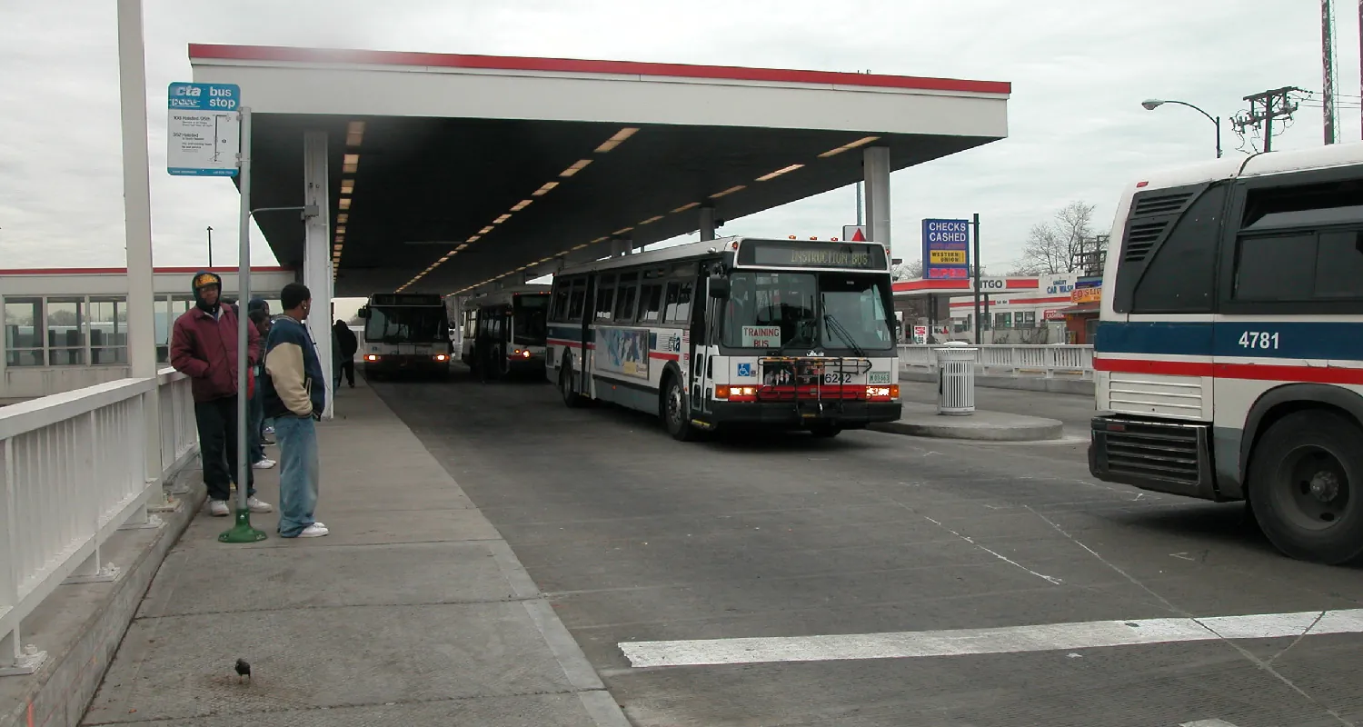 Two people stand outside at a bus stop. Three buses are parked at the stop and one bus is driving away.