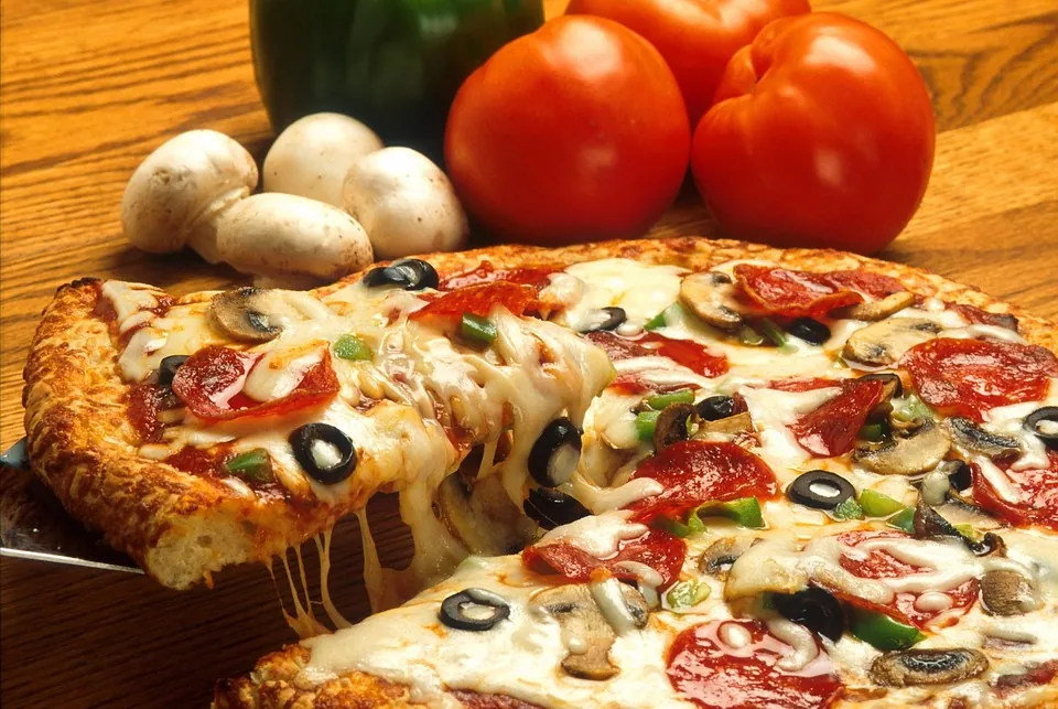 A delicious pizza slice with melted cheese stretching, topped with pepperoni, mushrooms, olives, and green peppers, alongside fresh vegetables on a wooden surface.
