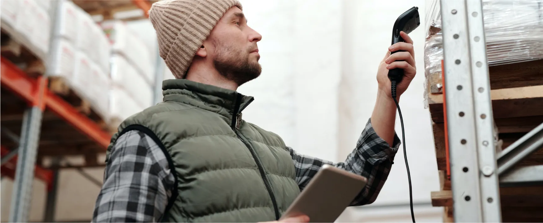 Image of a person holding a scanner and tablet in front of items on a shelf. Other shelves with items are visible in the back.