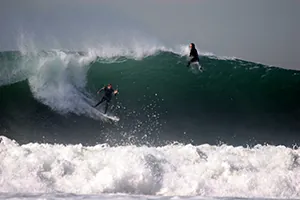 A surfer glides down a giant wave while another surfer watches from the wave’s crest.