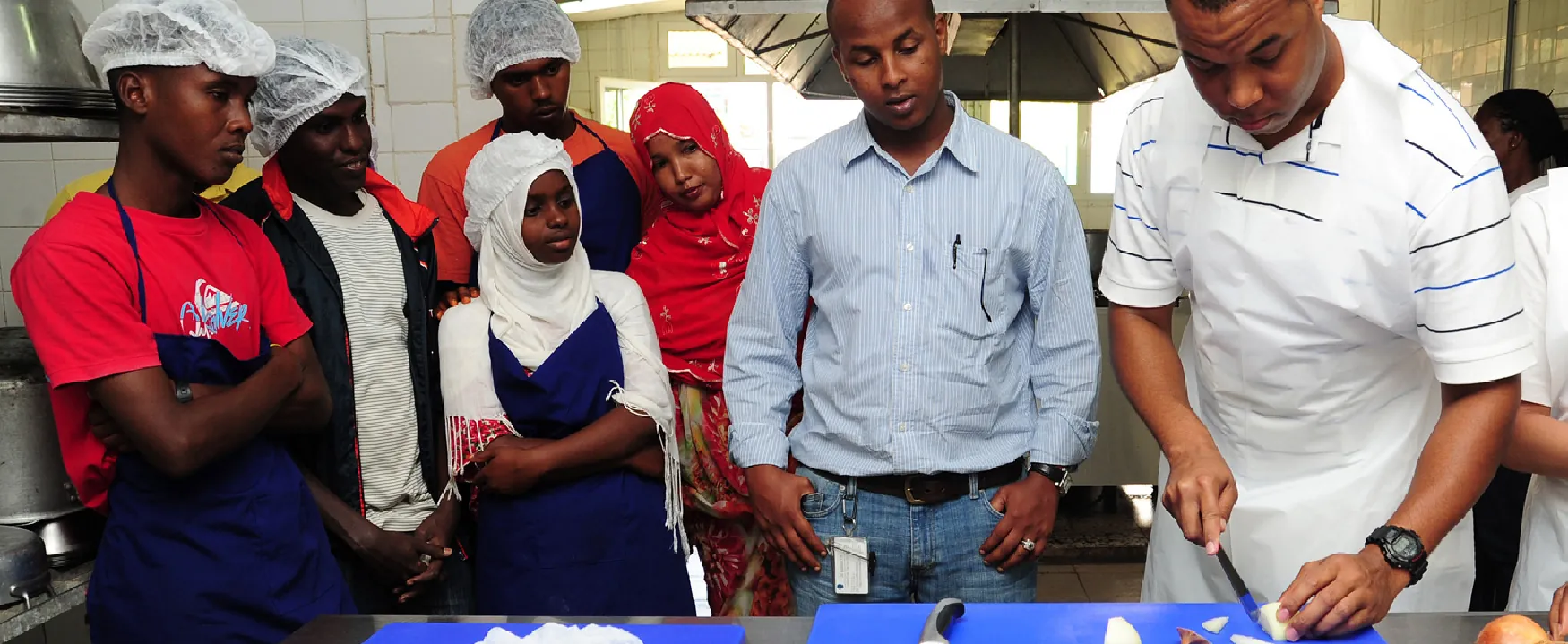 A group of people, some wearing hairnets and aprons, observe a person demonstrating how to chop food on a blue cutting board in a kitchen setting.