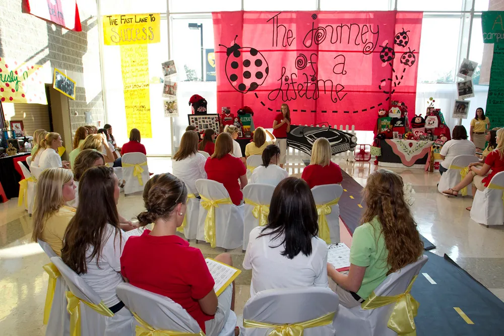 About a dozen young females are shown sitting in chairs at a sorority recruitment on campus.