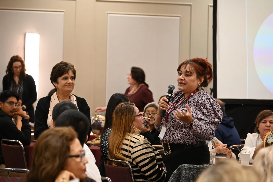 In a conference room, a person stands and speaks into a microphone. The speaker is surrounded by seated and standing people.